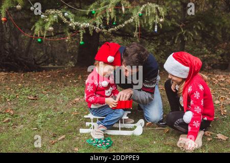 Père et deux filles ouvertes présente des boxes près de l'arbre de Noël en plein air dans la cour de la maison. Joyeux Noël et joyeuses fêtes Banque D'Images