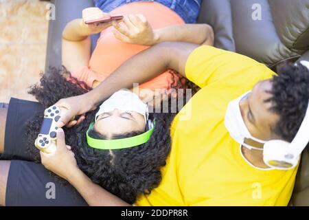 Garçon ayant le plaisir avec la console de jeux vidéo dans le temps de verrouillage, fille regardant le smartphone. Les jeunes du millénaire jouent en vacances dans la maison. Banque D'Images