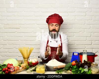 Le chef prépare le repas. Concept du processus de cuisson. Homme avec la barbe travaille avec la farine sur fond de mur de brique blanche. Faites cuire avec un visage curieux en uniforme assis près de la table avec des légumes, des pâtes et des ustensiles de cuisine Banque D'Images