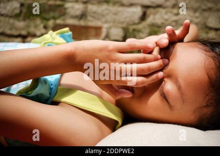 Ethnie d'Asie du Sud-est petite fille couché sur un banc, bronzant le matin à la maison. Les mains étant mises sur le visage pour se protéger de la lumière du soleil Banque D'Images