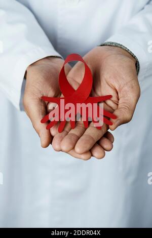 gros plan d'un médecin, en manteau blanc, avec un ruban de sensibilisation rouge, avec ses extrémités coupées en forme de mains, dans ses mains Banque D'Images