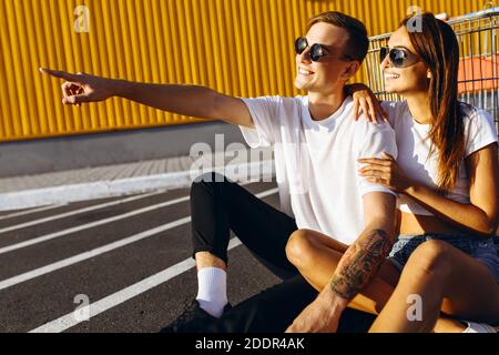 Un jeune homme et une fille, un gars pointe son doigt sur quelque chose au loin, assis sur l'asphalte dans la ville contre un mur jaune. Centre commercial Banque D'Images