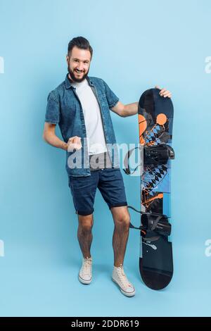 Un homme heureux avec une barbe , portant un short et une chemise , tient un snowboard et montre un geste de victoire et de succès, debout sur un bac bleu isolé Banque D'Images