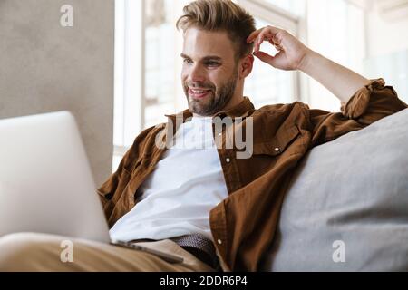 Beau homme souriant travaillant avec un ordinateur portable tout en étant assis sur un canapé à la maison Banque D'Images