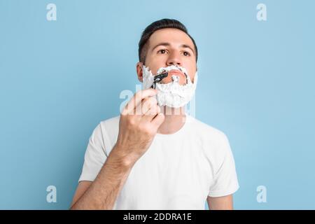 Un beau jeune homme avec une barbe se tient sur un un fond bleu avec de la mousse à raser sur son visage l'appareil photo et le rasage Banque D'Images