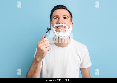 Un beau jeune homme avec une barbe se tient sur un un fond bleu avec de la mousse à raser sur son visage l'appareil photo et le rasage Banque D'Images