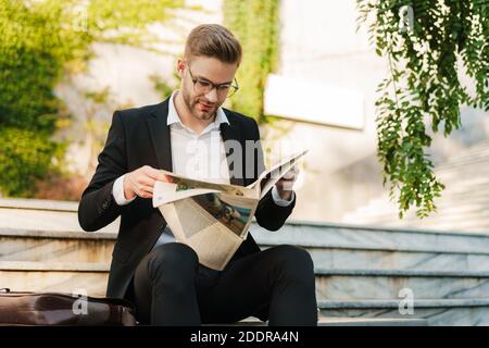 Beau homme d'affaires concentré lisant le journal tout en étant assis sur le banc près escaliers à l'extérieur Banque D'Images