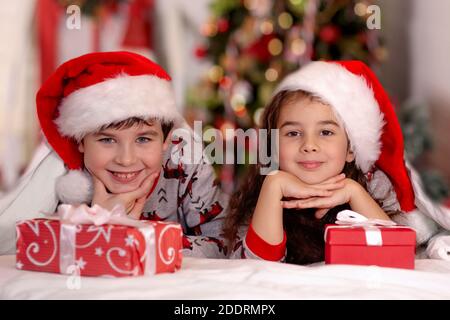 Deux enfants mignons, une fille et un garçon, en chapeaux du père noël, se câliner sur un lit blanc Banque D'Images