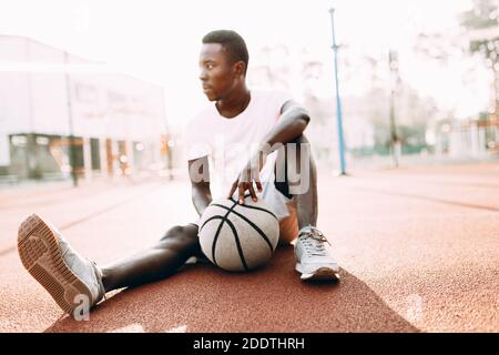 Jeune Afro-américain sportif reposant après un entraînement assis sur le terrain avec un joueur de basket-ball, un exercice matinal Banque D'Images