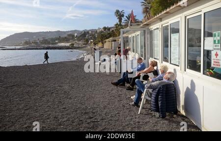 Personnes âgées assises sur des chaises en plastique, bains de soleil en Ligurie mer au milieu de l'hiver Banque D'Images