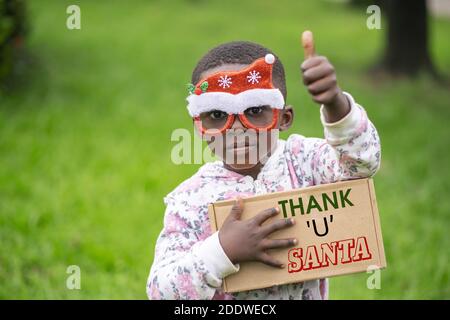 Un enfant africain avec des lunettes décoratives de Noël portant une plaque signalétique Avec l'écriture « Thank U Santa » Banque D'Images