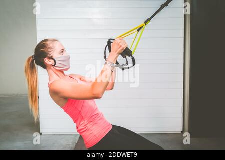 Jeune fille formant ses bras avec un masque de protection et des sangles TRx Fitness dans la salle de gym pendant la période de quarantaine pour la pandémie covid19 - femme faisant des pompes train Banque D'Images