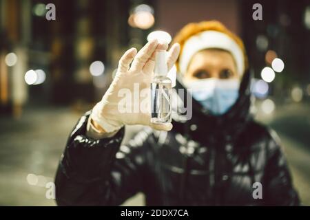 Femme mature avec masque facial et gants médicaux en caoutchouc tient antiseptique tout en se tenant contre la ville de nuit par temps froid. La femme âgée utilise la main Banque D'Images