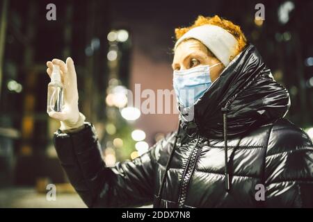 Protection coronavirus et covid 19. Antiseptique contre l'infection dans les mains de la femme mature dans la rue en soirée ville en hiver. Femme âgée en masque Banque D'Images