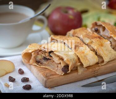 Une strudel aux pommes avec des raisins secs sur une planche en bois et sur fond de nappe blanche avec une tranche de pomme sur le côté, servi avec du café et de la crème - ho Banque D'Images
