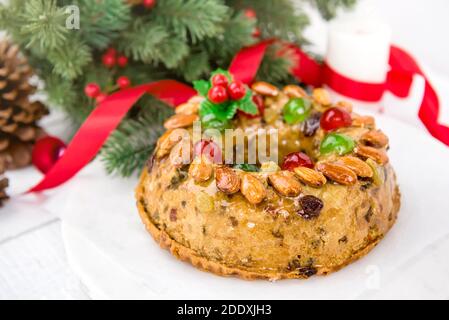 Gâteau de Noël rond vitrifié et coloré recouvert d'amandes et cerises de glace sur plaque blanche avec aiguilles de pin vert et ruban décoratif rouge Banque D'Images