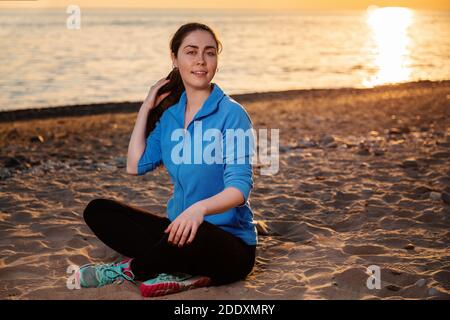 Bien-être et style de vie sportif. Une jeune femme souriante dans les vêtements de sport est assise à pieds croisés sur le sable et lissant ses cheveux. En arrière-plan t Banque D'Images