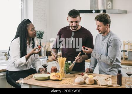 Femme afro-américaine avec deux hommes caucasiens cuisant une salade ensemble dans la cuisine à la maison le week-end Banque D'Images