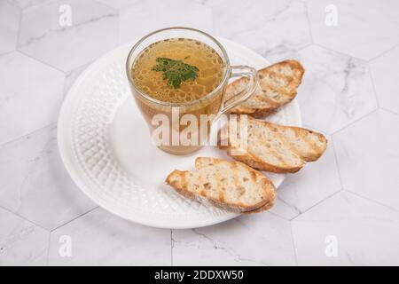 Bouillon de viande maison dans un verre tasse sur une plaque blanche avec des craquelins sur fond clair. Banque D'Images