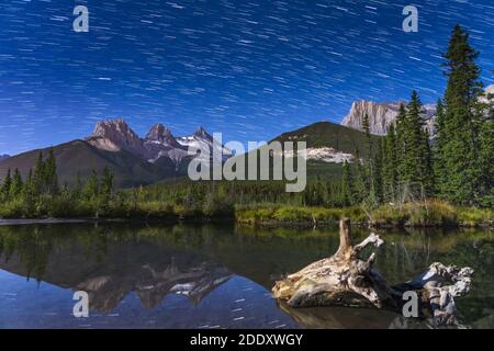 Pistes étoiles au-dessus de la montagne des trois Sœurs trio Peaks la nuit, ciel étoilé reflété dans la surface de l'eau du ruisseau. Magnifique paysage de la nation Banff Banque D'Images