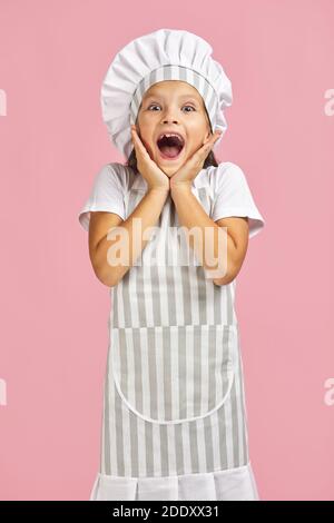 surprise petite fille surprise en chapeau de chef et tablier sur fond rose. Banque D'Images