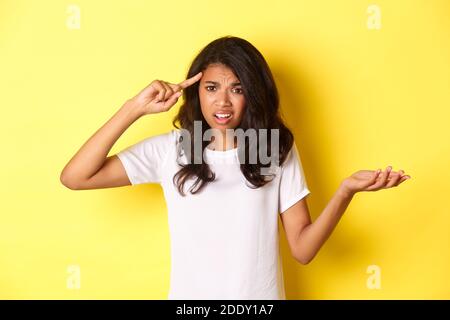 Portrait d'une jeune fille afro-américaine en colère et confuse, en colant quelqu'un pour avoir été stupide, pointant la tête et haussant les épaules, debout sur du jaune Banque D'Images