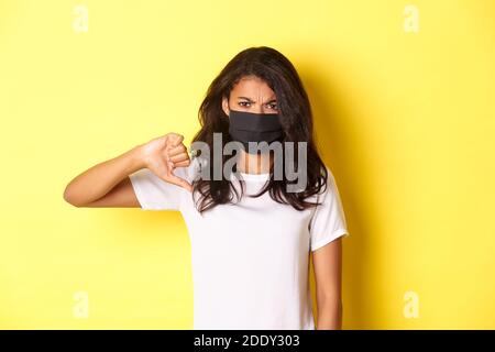 Concept de covid-19, distanciation sociale et style de vie. Image du modèle féminin afro-américain déçu, portant un masque noir, montrant des pouces Banque D'Images