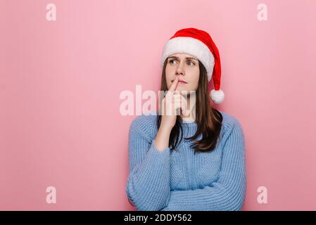 Une jeune femme confuse dans un chapeau de Noël avec une expression perplexe, se répand les mains avec un look perplexe, se dresse perplexe, isolée sur fond de studio rose Banque D'Images