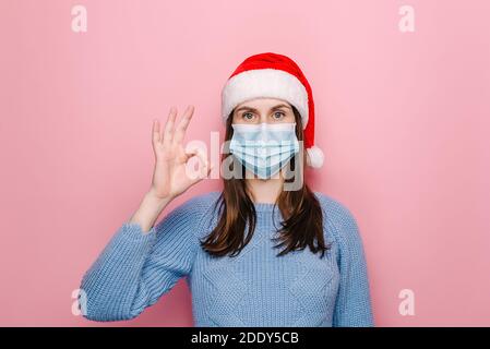 Belle jeune femme en chapeau rouge de Noël montrant le signe de geste ok, porte un masque médical de protection pour ne pas infecter d'autres personnes, vêtu chandail d'hiver Banque D'Images