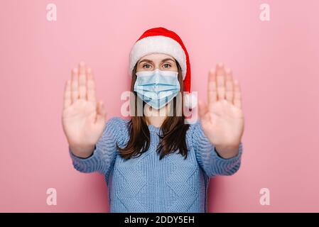 Une jeune femme sérieuse en masque médical tire les mains vers l'appareil photo en geste d'arrêt, montre la limite, porte un chandail bleu et chapeau de Noël, isolé sur le mur rose Banque D'Images