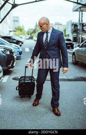 Homme d'affaires chauve marchant sur le parking de l'aéroport avec des valises et des voitures garées. Homme d'affaires en costume et valise lors d'un voyage d'affaires. Homme marchant Banque D'Images