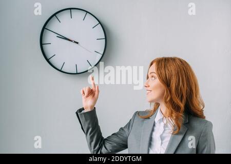 Femme d'affaires souriante, à tête rouge, pointant du doigt et regardant le mur horloge sur gris Banque D'Images