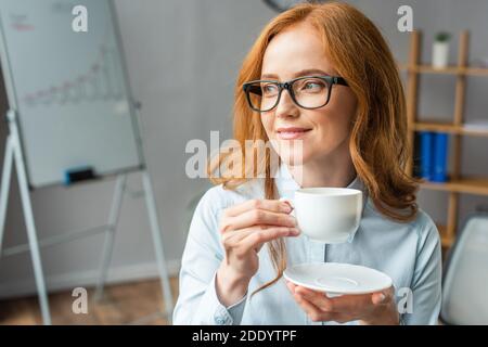 Femme d'affaires souriante à tête rouge qui regarde loin, tout en tenant une tasse à café et une soucoupe avec un tableau de conférence flou sur l'arrière-plan Banque D'Images