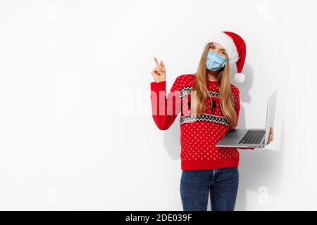 Jeune femme heureuse portant le chapeau du père noël, chandail de noël, masque de protection médicale sur le visage, en utilisant un ordinateur portable et le doigt pointant vers l'espace de copie, sur wh Banque D'Images