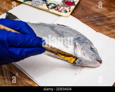 Master class pour le dessin de poissons en technique Monotype - main gantée Peint le poisson Orata cru avec de la peinture acrylique argentée sur le papier serviette sur la table recouverte de Banque D'Images