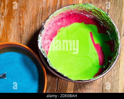 cuisiner un gâteau sucré à la maison - vue de dessus moule à charnière avec mélange de creux liquides verts et rouges et un bol de pâte bleue sur une ancienne table en bois Banque D'Images