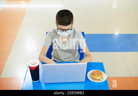 Un adolescent portant un masque facial utilise un ordinateur portable à la restauration rapide restaurant pendant le déjeuner Banque D'Images