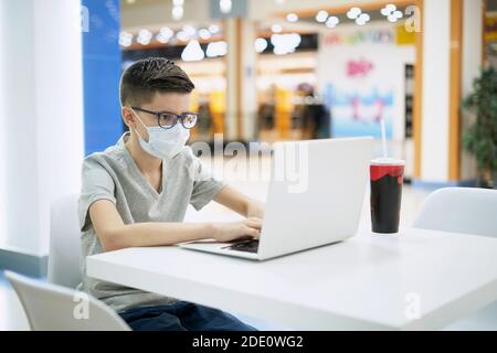 Un adolescent portant un masque facial utilise un ordinateur portable lorsqu'il est assis à l'aire de restauration Banque D'Images