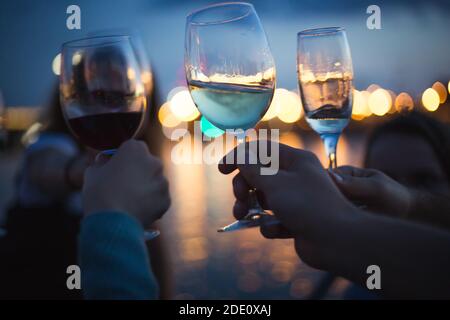 une fête à l'extérieur, célébrant et élevant des verres avec du vin contre le ciel bleu de nuit et les lumières de la ville jaune, le bokeh agréable et les réflexions de lumières dans l'eau Banque D'Images