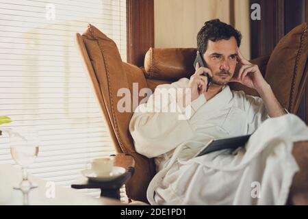 Un homme dans un peignoir blanc a un appel de la chambre d'hôtel. Assise dans un confortable fauteuil plus confortable avec une tablette sur les genoux Banque D'Images