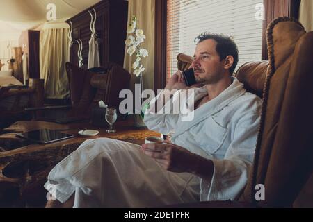 Un homme dans un peignoir blanc a un appel de la chambre d'hôtel. Assise dans un confortable fauteuil plus confortable avec une tablette sur les genoux et un café sur le t Banque D'Images