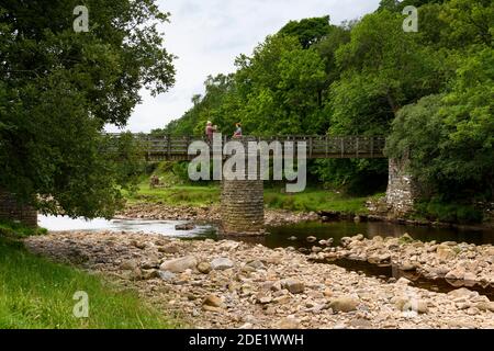 Passerelle au-dessus de la rivière Swale (campagne pittoresque, eau peu profonde, rochers de la rivière, 2 personnes prenant des photos) - rampes Holme Bridge, Yorkshire Dales, Royaume-Uni. Banque D'Images