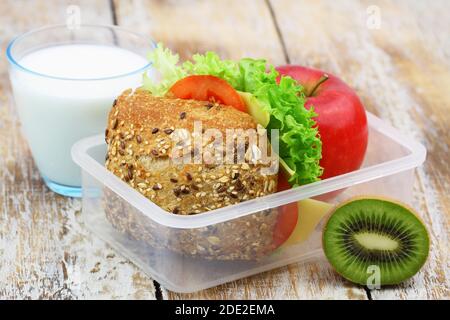 Boîte à lunch nutritive contenant un rouleau de fromage brun avec de la laitue et de la tomate, de la pomme, des kiwis et un verre de lait Banque D'Images