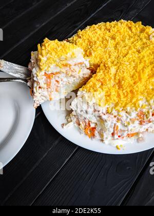 Salade russe maison Mimosa avec des pommes de terre, du poisson, des carottes, de la mayonnaise et râpez des jaunes d'œufs dans une assiette sur une table en bois sombre. Une tranche a été coupée et moi Banque D'Images