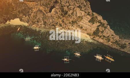 Bateaux passagers traditionnels des Philippines au coucher du soleil, vue aérienne. Transport d'eau à la baie océanique avec plage de sable côte de l'île de montagne de Palawan, archipel de Visayas. Paysage marin bleu foncé spectaculaire Banque D'Images