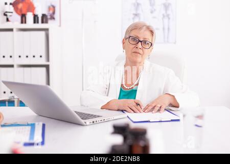 Femme mature médecin portant un blouse de laboratoire dans la chambre d'hôpital tout en utilisant un ordinateur portable. Médecin utilisant le bloc-notes dans le milieu de travail de la clinique , confiant, expertise, médecine. Banque D'Images