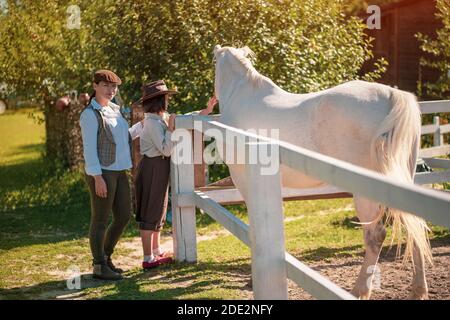 Maman et fille dans les vêtements des fermiers vintage stand près du paddock avec un beau cheval. La fille est à la course d'un cheval, maman regarde et sourit à la Banque D'Images