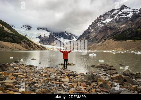 Vue arrière du jeune homme en veste rouge debout à bras ouverts par Laguna Torre, Parc national Los Glaciares, El Chalten, Patagonie, Argentine Banque D'Images