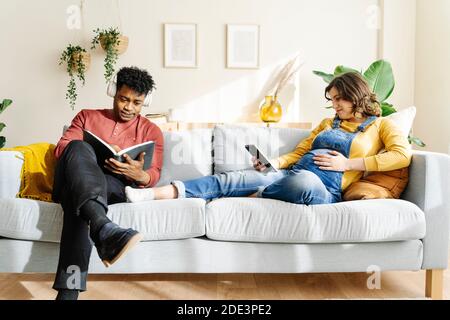 Mari écoutant de la musique et lisant un livre avec sa femme enceinte qui regarde une tablette sur un canapé. Concept de couple interracial Banque D'Images