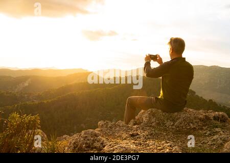 Un homme photographiant le coucher du soleil avec son mobile assis sur un rocher au sommet de la montagne. Concept de la nature Banque D'Images